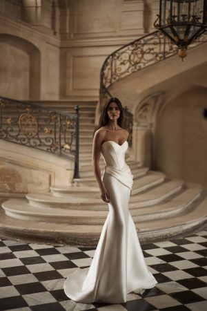 Diane by Milla Nova. Sophisticated mermaid gown in mikado with masterful drapery accentuating the hips. Featuring a heart-shaped neckline with integrated cups, a structured corset, lace-up closure at the back, and a form-fitting mermaid skirt with draping on the hips and a short train.