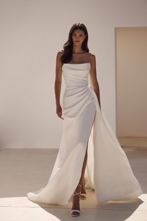 Sandal by Milla Nova. A Refined fitted wedding gown cut from satin fabric. Featuring a geometric neckline, lace-up or zipper closure on the back, and a beautifully draped waist that gracefully transitions into a gorgeous layering skirt with a delicate train.