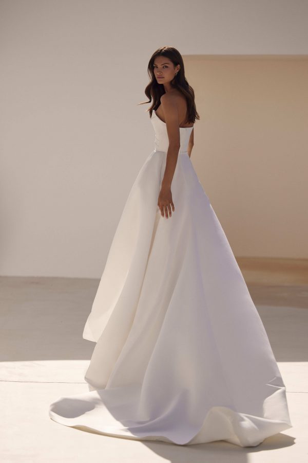 Sandal by Milla Nova. A Refined fitted wedding gown cut from satin fabric. Featuring a geometric neckline, lace-up or zipper closure on the back, and a beautifully draped waist that gracefully transitions into a gorgeous layering skirt with a delicate train.