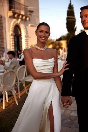 Lancaster by Milla Nova. A Timeless A-line classic wedding gown with draping. The sleeveless design features a corset with a cinched waist, a draped neckline, a zipline on the back decorated with buttons, and a flowy full skirt with a long train and side slit.