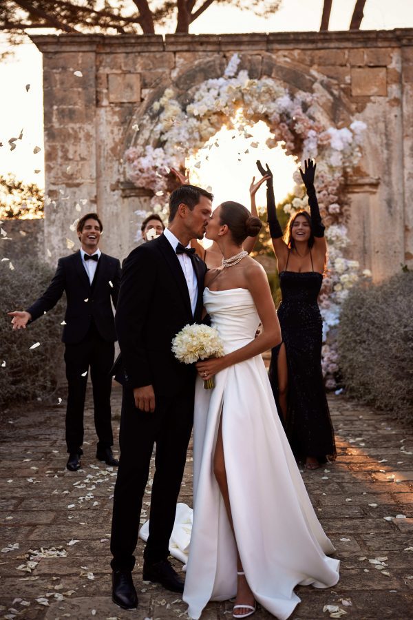 Lancaster by Milla Nova. A Timeless A-line classic wedding gown with draping. The sleeveless design features a corset with a cinched waist, a draped neckline, a zipline on the back decorated with buttons, and a flowy full skirt with a long train and side slit.