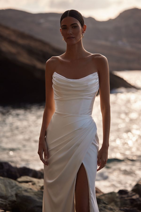 Bellissima by Milla Nova. A Glamorous and sleek satin A-line wedding gown with elegant draping. The sleeveless design features a structured corset with visible boning, a sharp artfully draped neckline, and a fitted maxi skirt with a daring thigh-high slit. Side draping accentuates the silhouette, leading to a daring side slit and finishing with a subtle train. Can be complimented by a chic satin off-the-shoulder bolero Bellissima with long sleeves. You can also choose a detachable train to extend the dress.