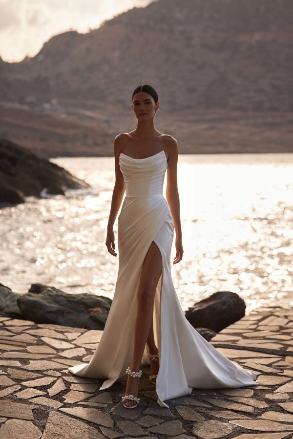Bellissima by Milla Nova. A Glamorous and sleek satin A-line wedding gown with elegant draping. The sleeveless design features a structured corset with visible boning, a sharp artfully draped neckline, and a fitted maxi skirt with a daring thigh-high slit. Side draping accentuates the silhouette, leading to a daring side slit and finishing with a subtle train. Can be complimented by a chic satin off-the-shoulder bolero Bellissima with long sleeves. You can also choose a detachable train to extend the dress.