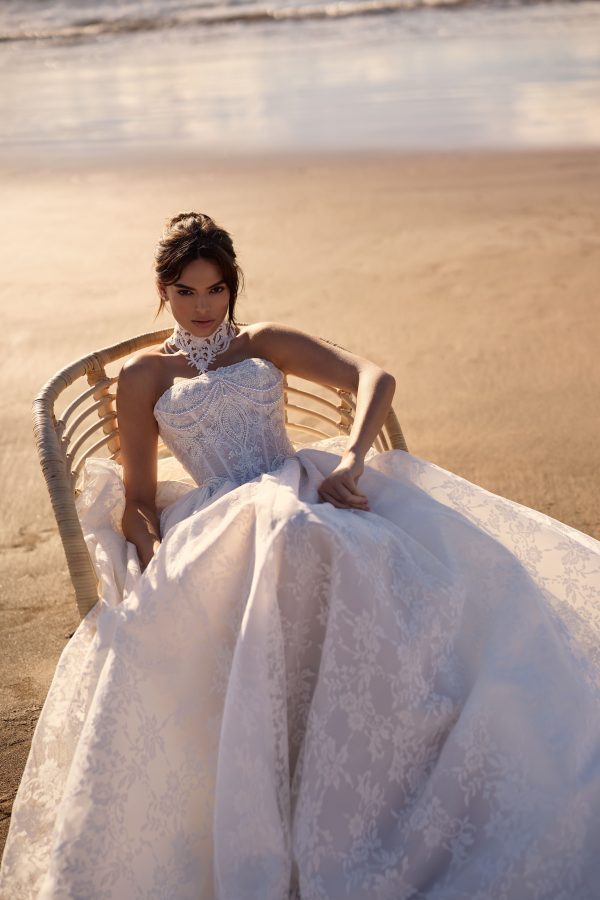 Monica by Milla Nova. A Vintage-inspired exquisite wedding gown cut in a princess silhouette from delicate chantilly lace. Featuring a straight neckline, a structured bodice with built-in cups and visible boning that lengthens the silhouette, a zip closure, and a voluminous lace skirt with a train that flares out from the corset. This ballgown features a baroque-inspired ornament embellished with sequins. Includes detachable pearl jewelry for versatile styling — wear it on the corset or as a necklace. For a complete and romantic look, the gown can be styled with an exquisite lace Monica choker and ruched lace Monica cuffs. The dress includes detachable off-the-shoulder sleeves.