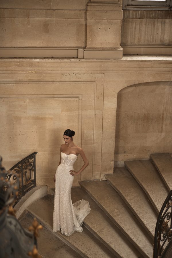 Aminata by Milla Nova. A Gleaming fitted wedding gown cut from lace in a mermaid silhouette, adorned with dozens of sequins. Featuring a heart-shaped neckline, a boned corset with built-in cups, a zip closure on the back, and a fitted maxi skirt with a short train. The gown can be elevated with a stunning satin illusion cover dress standing out with a ruched bodice and an asymmetrical ruched mini skirt with a long train.