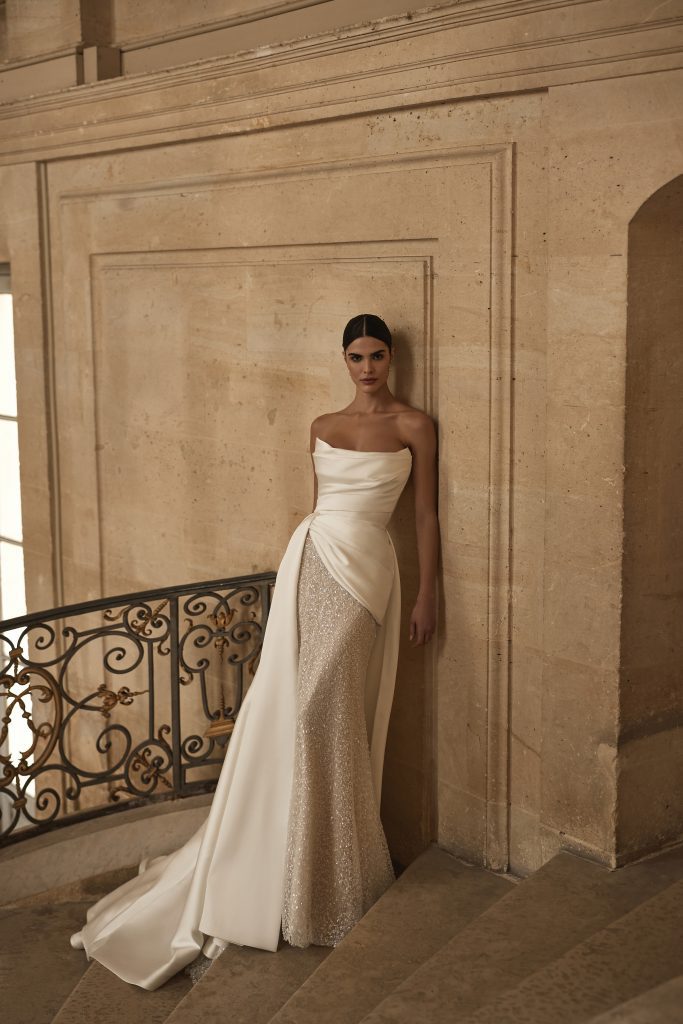 Gleaming fitted wedding gown cut from lace in a mermaid silhouette, adorned with dozens of sequins. Featuring a heart-shaped neckline, a boned corset with built-in cups, a zip closure on the back, and a fitted maxi skirt with a short train. The gown can be elevated with a stunning satin illusion cover dress standing out with a ruched bodice and an asymmetrical ruched mini skirt with a long train.