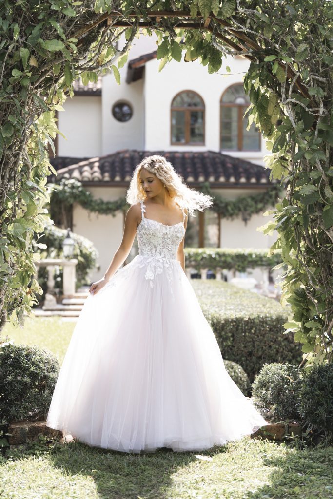 Betty by Allure Romance. Made for a grand entrance, this airy ballgown balances volume with delicate detail. A scoop neckline and thin lace-appliquéd straps frame the subtle sheer corset bodice, where exposed boning adds just the right touch of structure. Dimensional lace and tonal beading trail over soft layers of tulle, while a natural waist and floor-length hem complete the look.