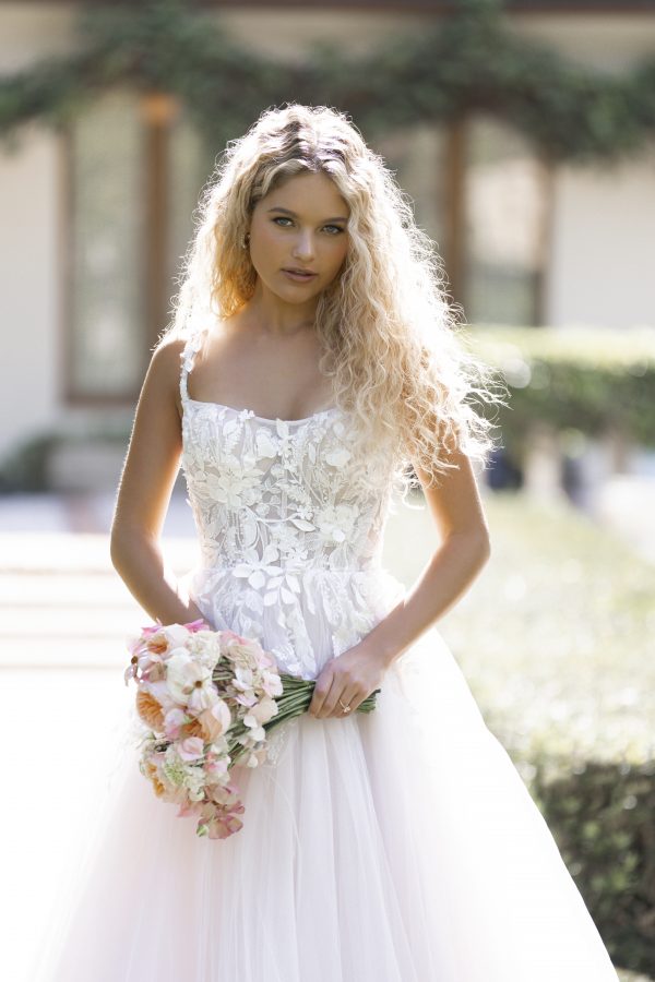 Betty by Allure Romance. Made for a grand entrance, this airy ballgown balances volume with delicate detail. A scoop neckline and thin lace-appliquéd straps frame the subtle sheer corset bodice, where exposed boning adds just the right touch of structure. Dimensional lace and tonal beading trail over soft layers of tulle, while a natural waist and floor-length hem complete the look.