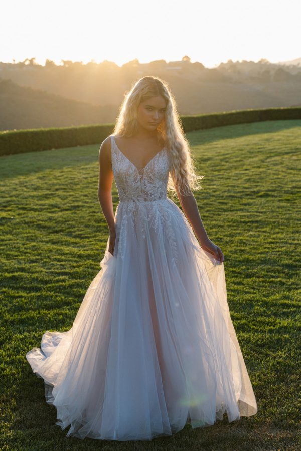Radiant and graceful, this A-line gown features pearl, beaded, and embroidered lace appliques that glisten over layers of sparkle tulle. The V-neckline and slender straps create a flattering, open frame, while the natural waist flows into a soft, sweeping skirt. With its gentle shimmer and intricate lacework, this gown captures a beautifully romantic sensibility with just the right touch of light-catching detail.